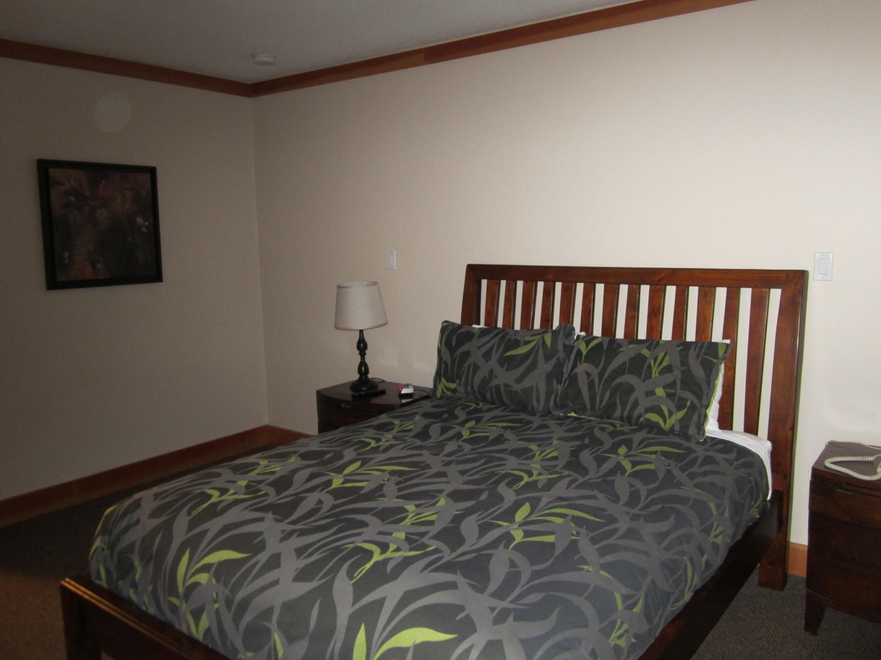 A neatly made bed with a patterned bedspread, a wooden headboard, a bedside table with a lamp, and a framed picture on the wall.