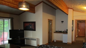 A bedroom with a bed, a TV, a painting, a kitchenette, and a door leading to another room.