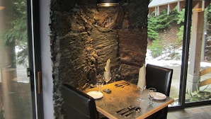 A dining area with a stone wall, a table set with cutlery and glasses, and a view of the outdoors through large windows.