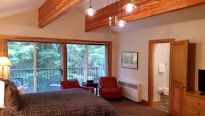 A bedroom with a large bed, a red chair, a table, and a view of a balcony with a railing. There is a door to the right and a framed picture on the wall.