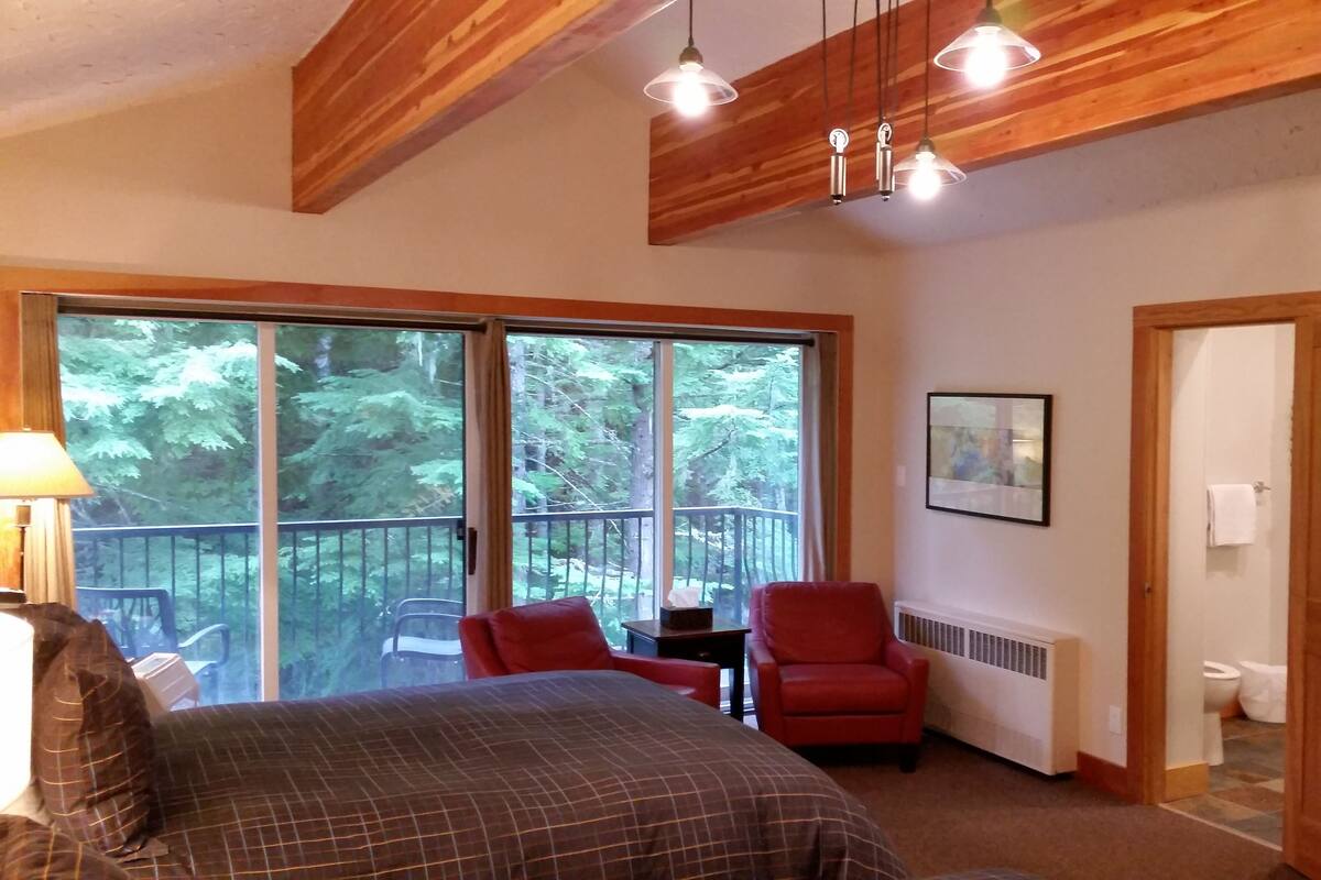 A bedroom with a large bed, a red chair, a table, and a view of a balcony with a railing. There is a door to the right and a framed picture on the wall.