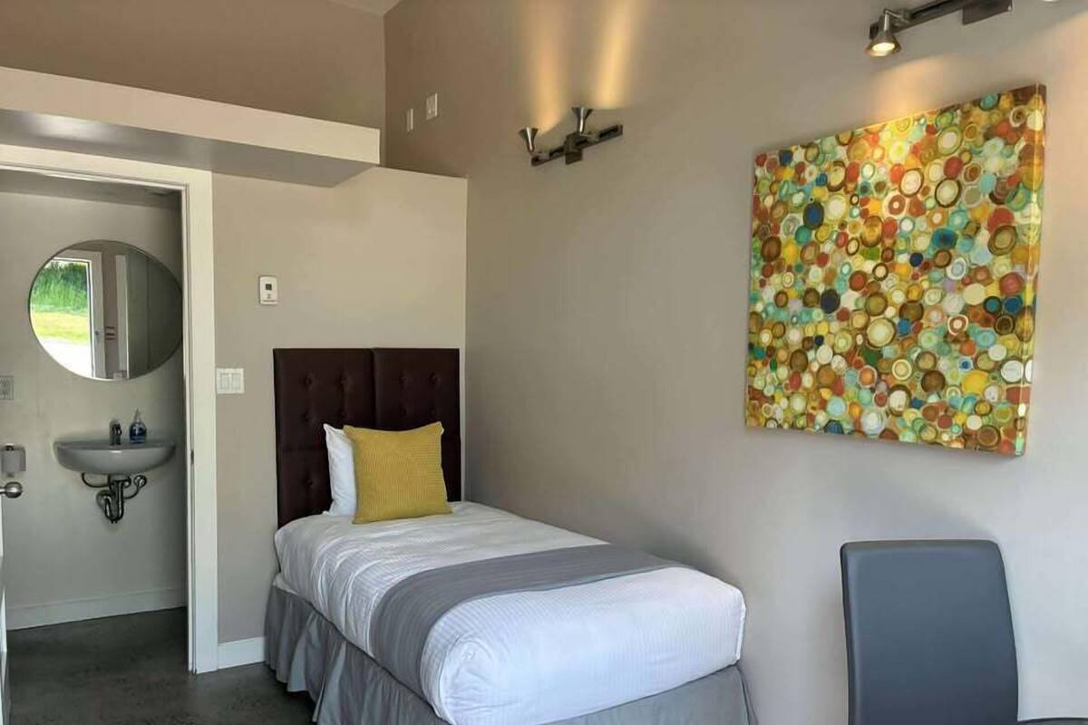 Une chambre à coucher comprenant un lit, une table de chevet, un miroir, un lavabo et un tableau abstrait aux couleurs vives accroché au mur.
