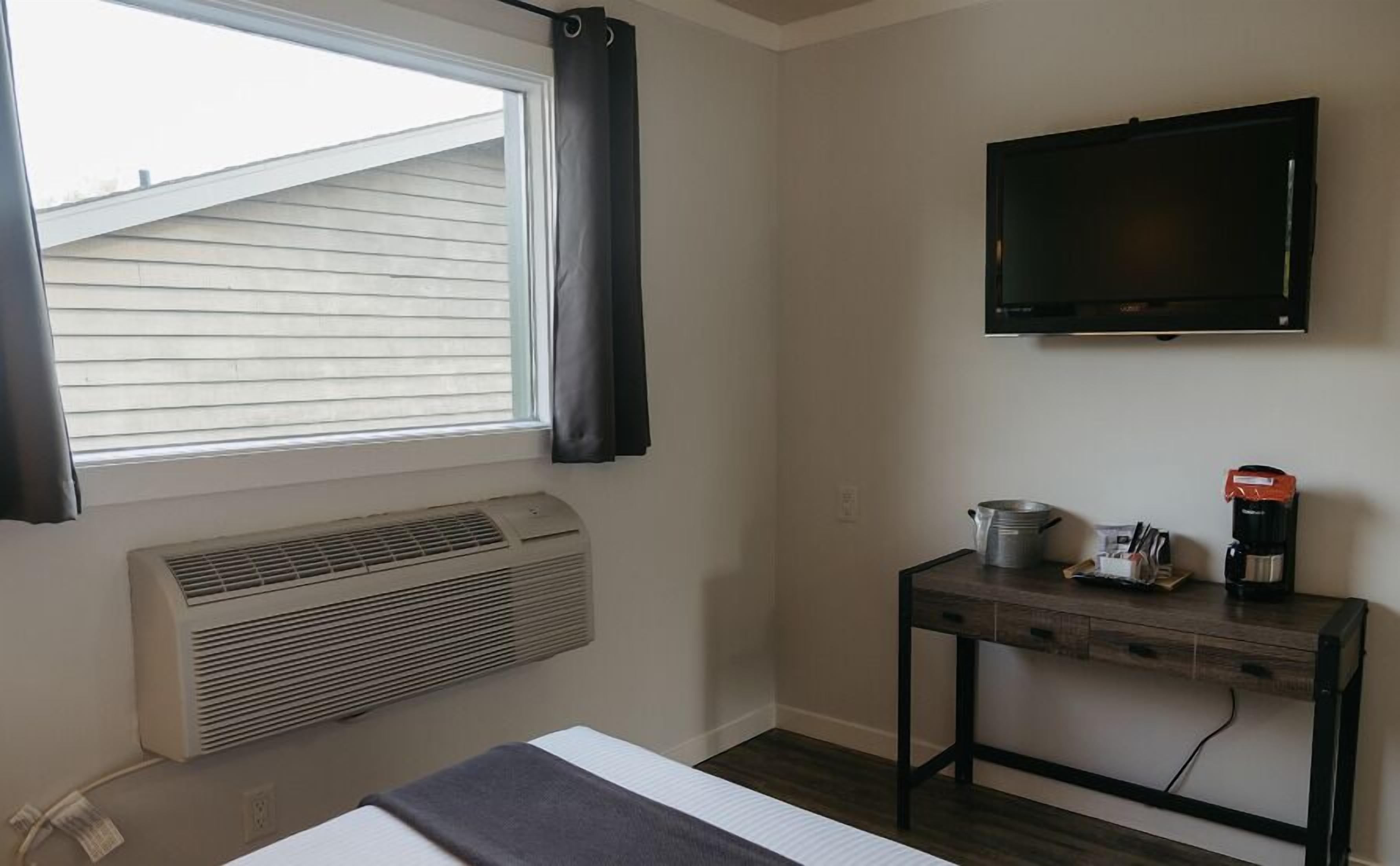 Una habitación con cama, ventana con cortinas, televisor montado en la pared, calentador, una mesita con utensilios de cocina y una cafetera.