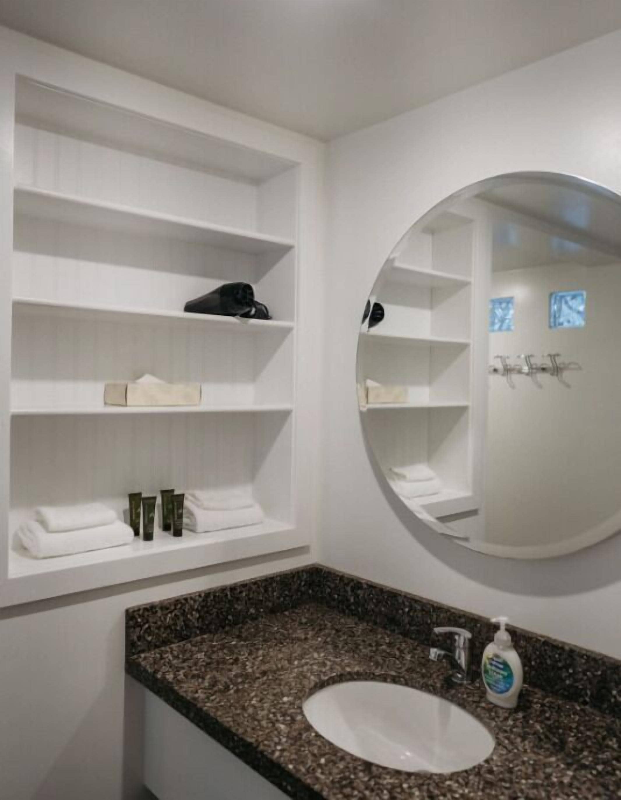 Une salle de bain dotée d’un comptoir en granit, d’un lavabo blanc et d’étagères où sont rangés les produits de toilette.