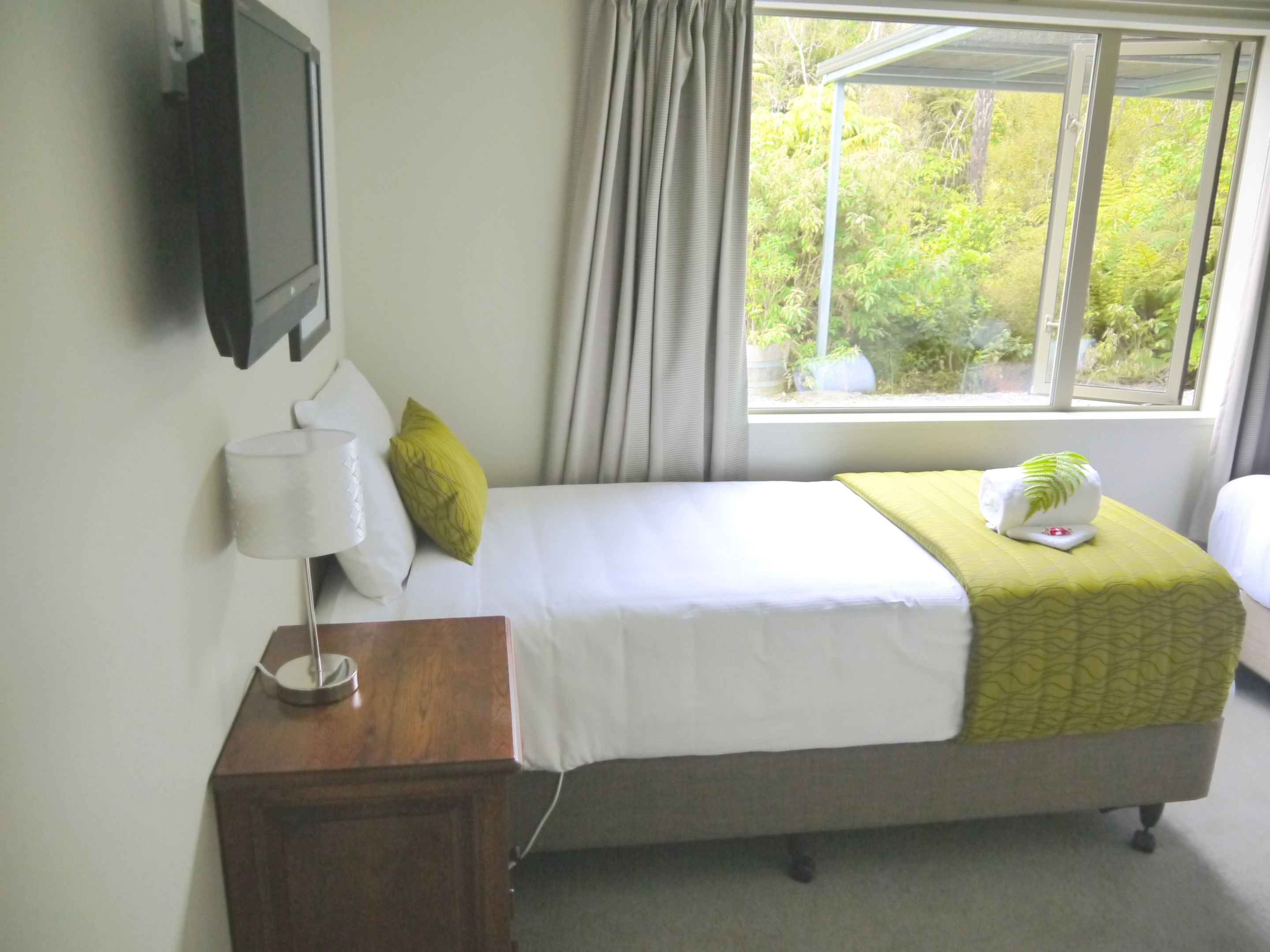 Habitación de hotel con cama, mesita de noche con lámpara, ventana con cortinas y vistas al exterior.