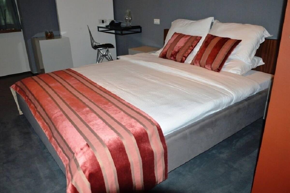 Un lit bien fait, recouvert d’une couvre-lit à rayures, une table de chevet avec une lampe et une chaise dans une chambre moderne aux murs gris.