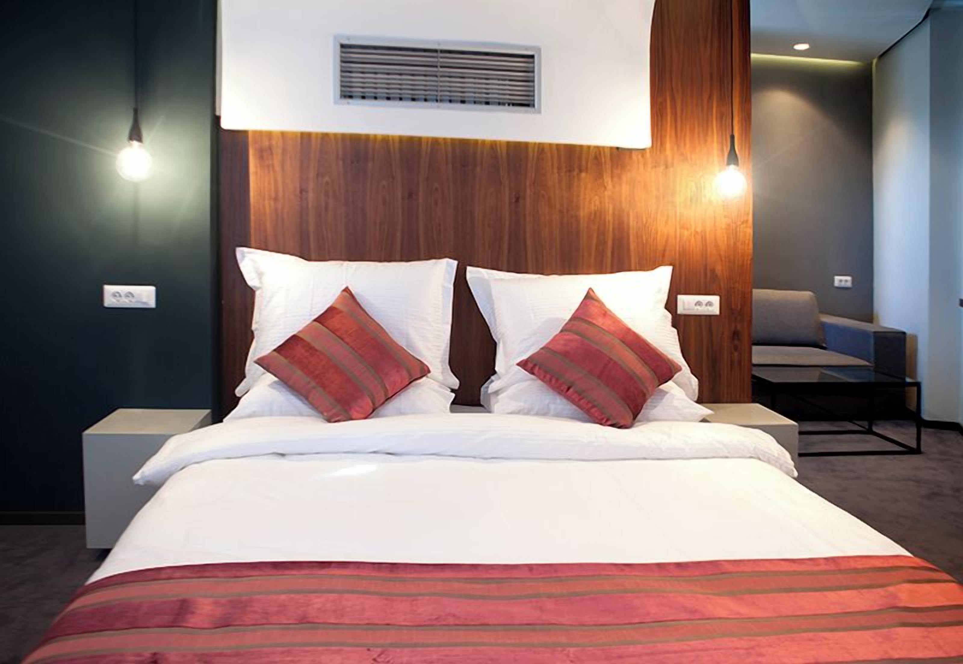 A hotel room with a bed, two bedside lamps, a chair, and a wall-mounted air vent.