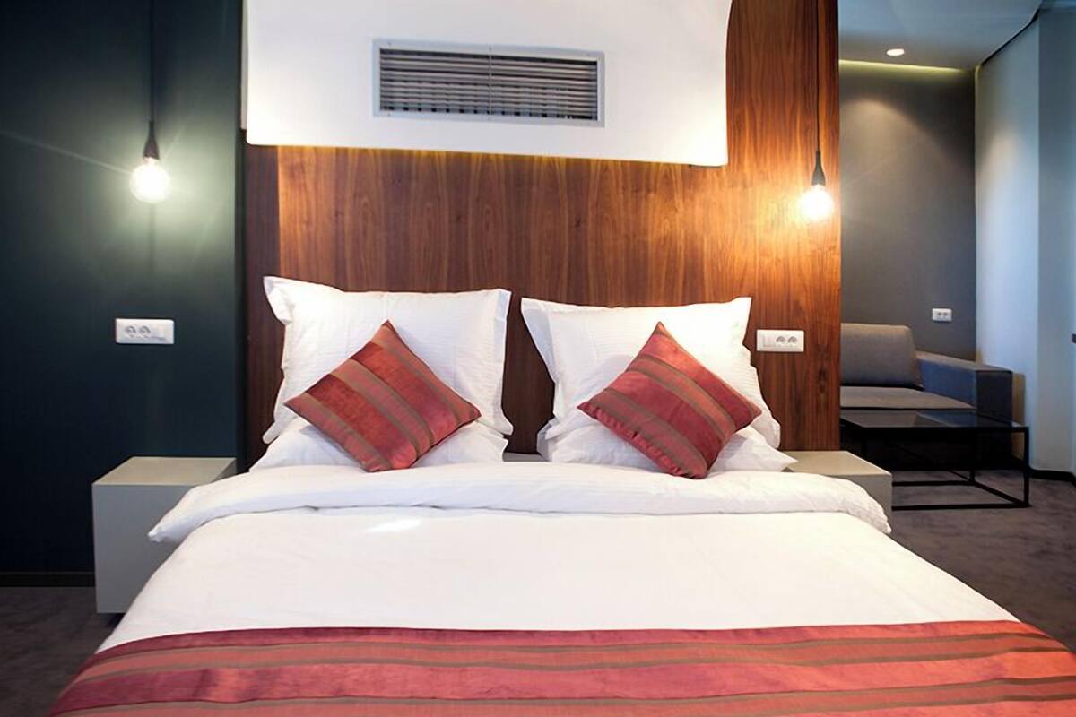 Une chambre d’hôtel avec un lit, deux lampes de chevet, une chaise et une bouche d’aération fixée au mur.