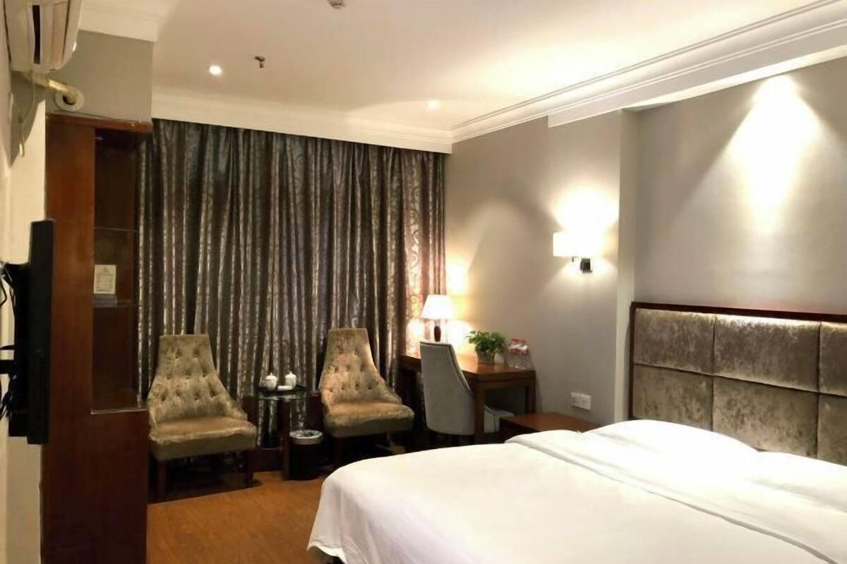 Une chambre d’hôtel avec un grand lit, deux fauteuils, un bureau avec une lampe et un rideau.