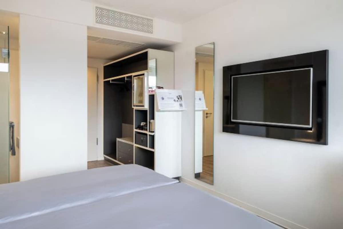 Habitación de hotel con una cama, un armario, un televisor montado en la pared y un pequeño escritorio con una silla.