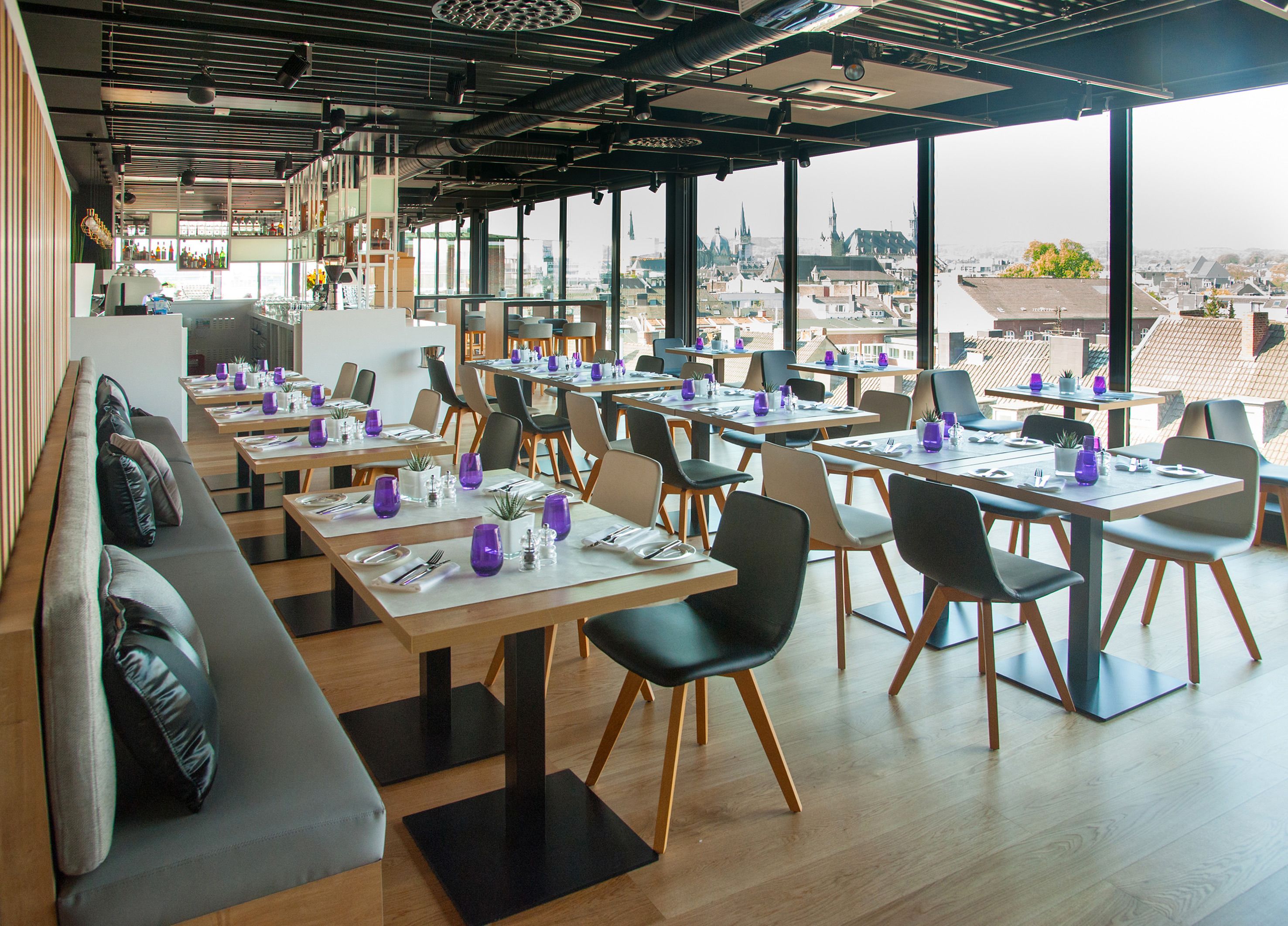 Un restaurante moderno con mesas y sillas altas, una zona de bar y grandes ventanales con vistas a la ciudad.