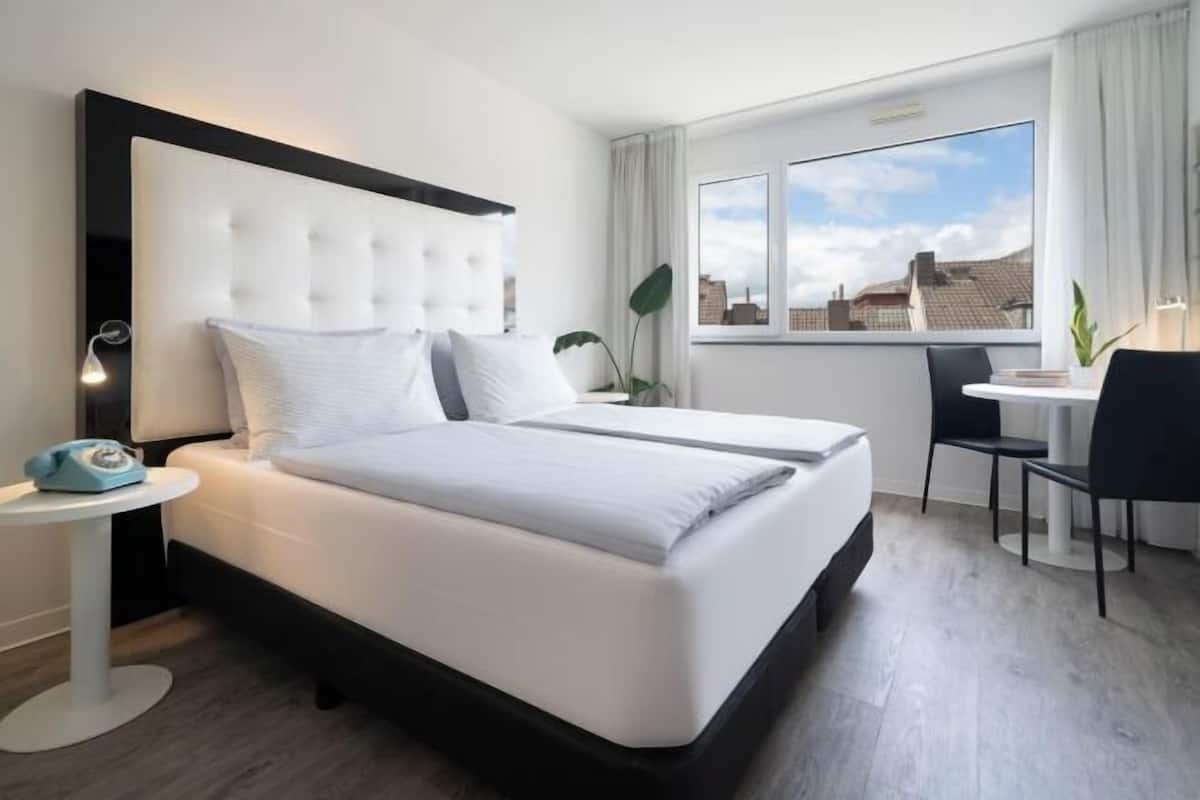 A modern hotel room with a large bed, a small desk, a chair, and a window with a city view.