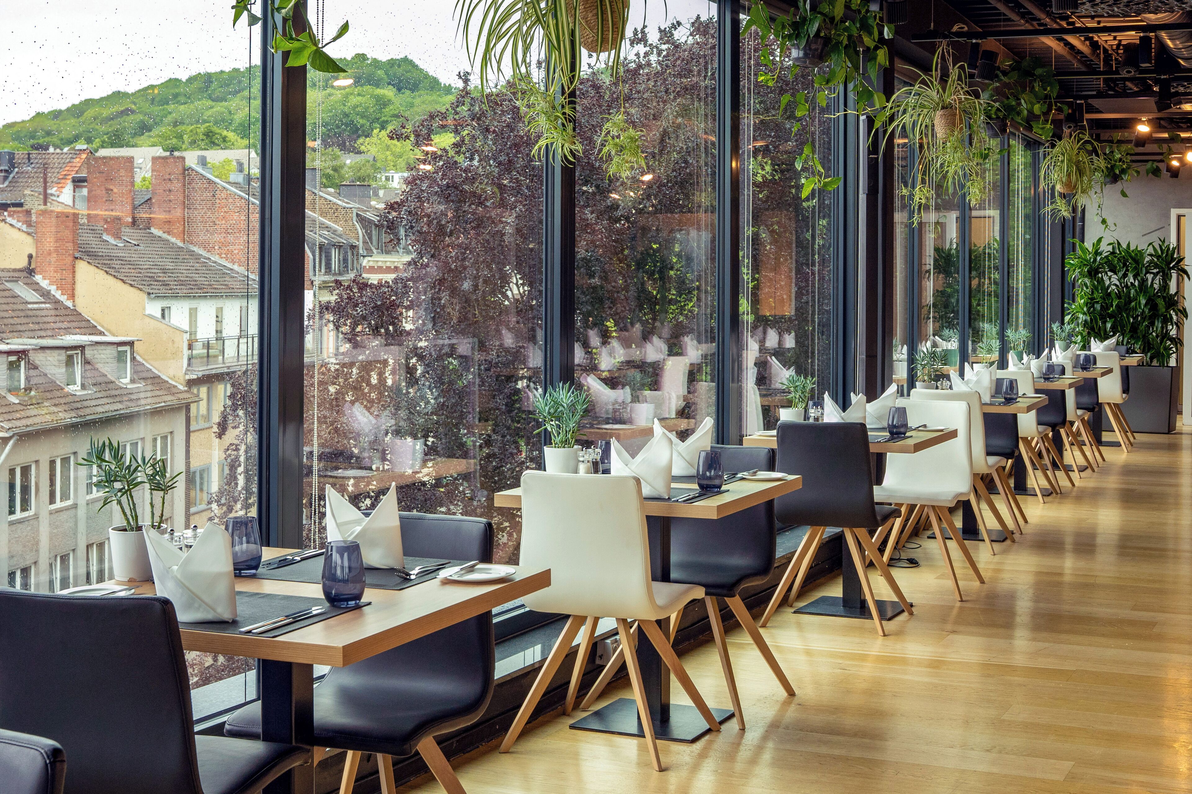 A modern restaurant with large windows, wooden tables, black chairs, and potted plants.