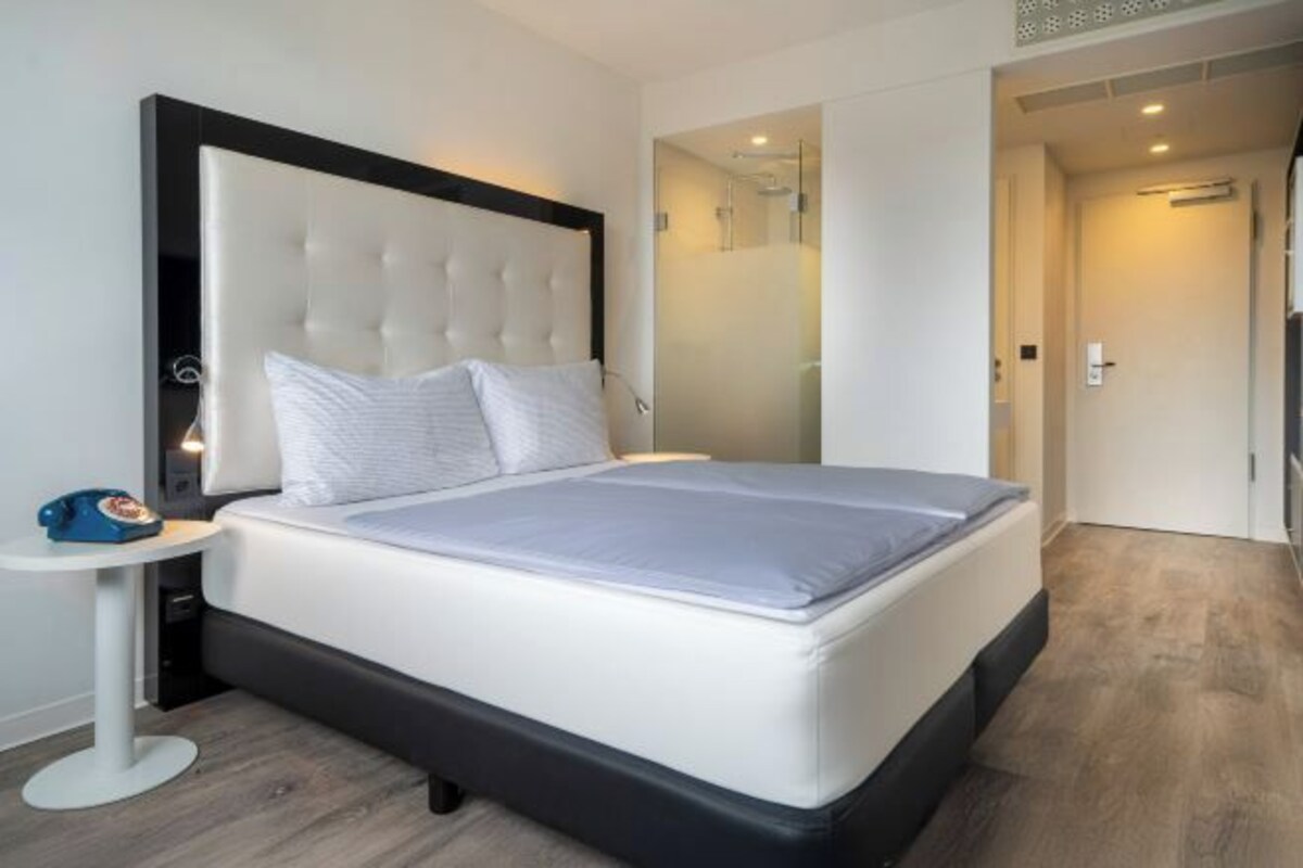 Una habitación de hotel moderna con una cama grande, una mesita de noche y un box de ducha con paredes de vidrio.