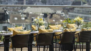 Comedor con mesas dispuestas para dos, con servilletas amarillas y cristalería, con vistas a un paisaje urbano.