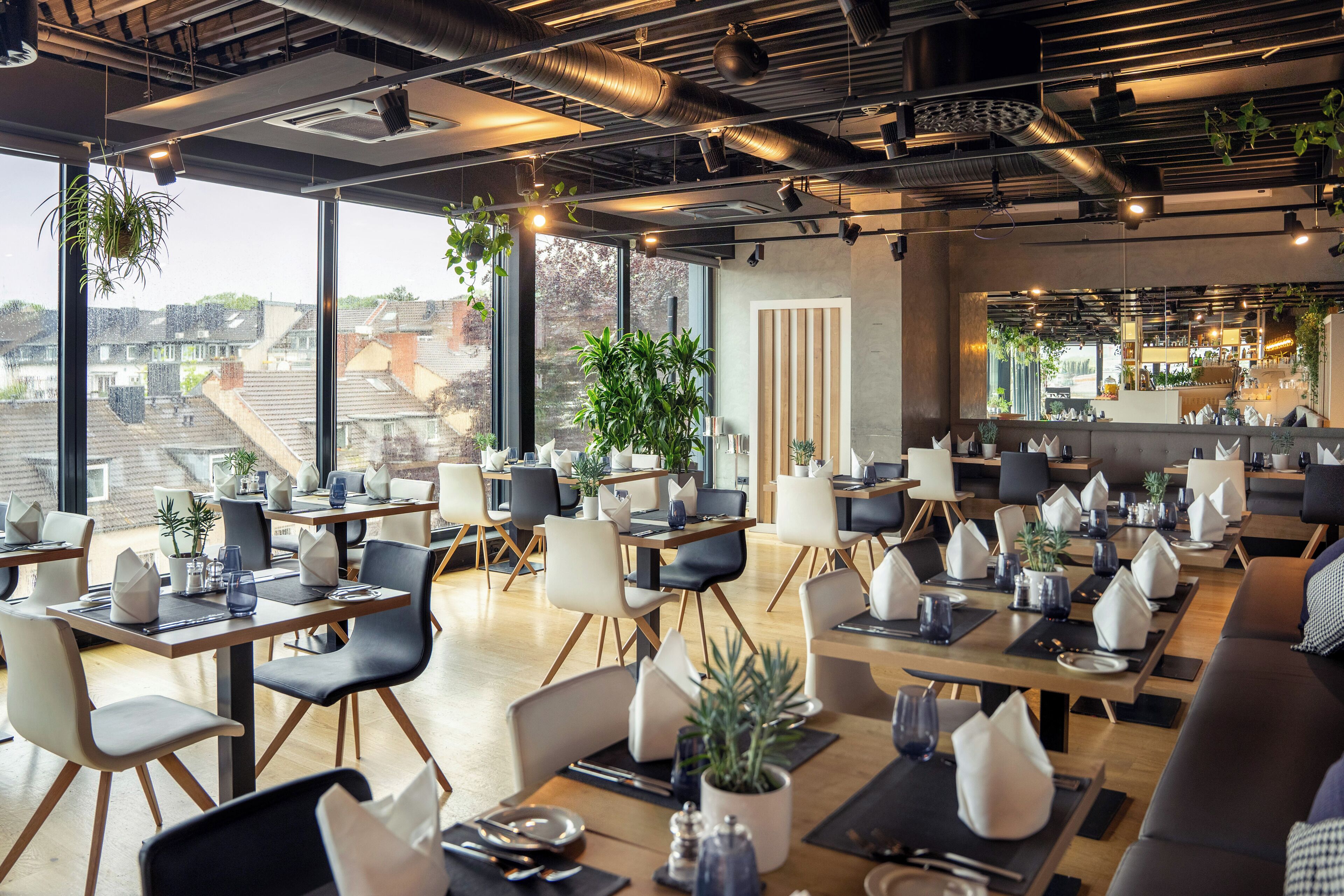 Ein modernes Restaurant mit hohen Decken, freiliegenden Lüftungsrohren und großen Fenstern mit Blick auf die Stadt. Tische sind ordentlich mit weißen Servietten und Topfpflanzen angerichtet.
