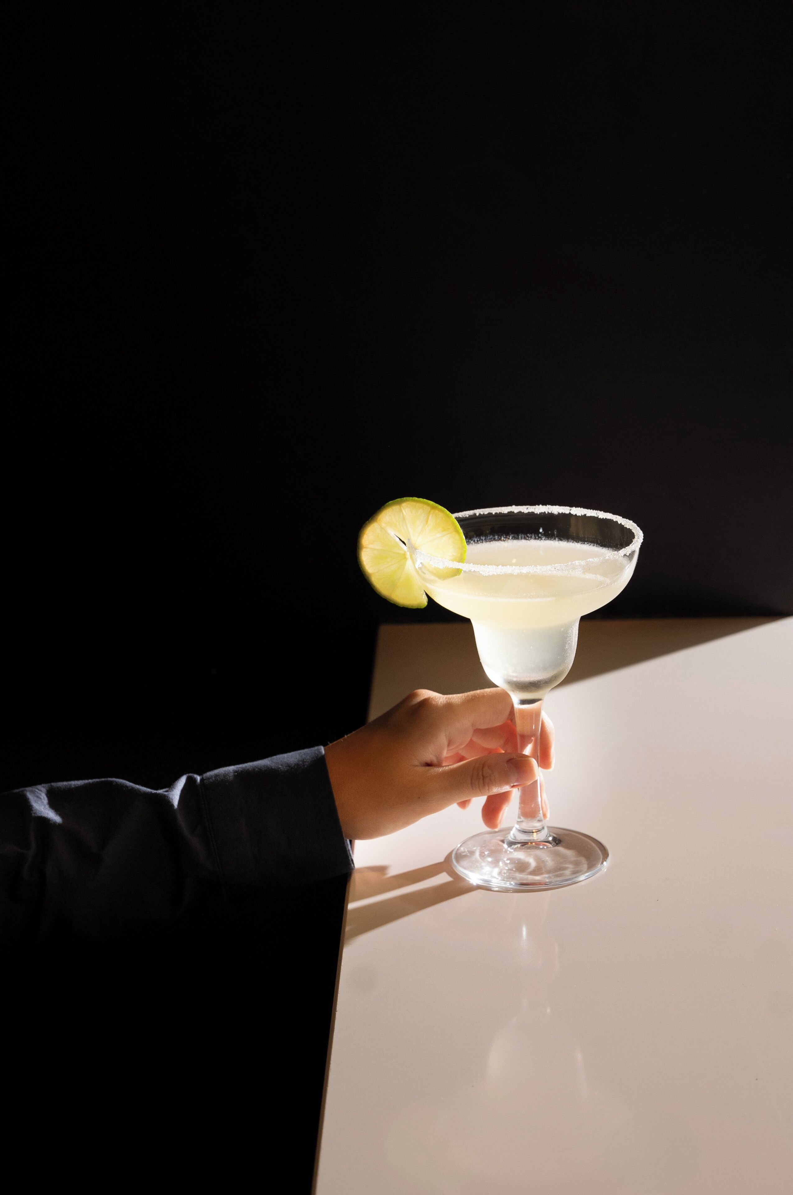 Eine Hand hält ein Cocktailglas mit Limettenscheibe auf einem Tisch.