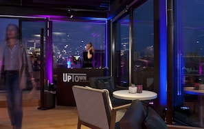 A modern interior with a bar area, a seating area with a round table, and large windows offering a city view.