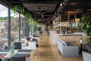 Un restaurante moderno con una barra larga, techos altos y amplios ventanales con vistas a un paisaje urbano.