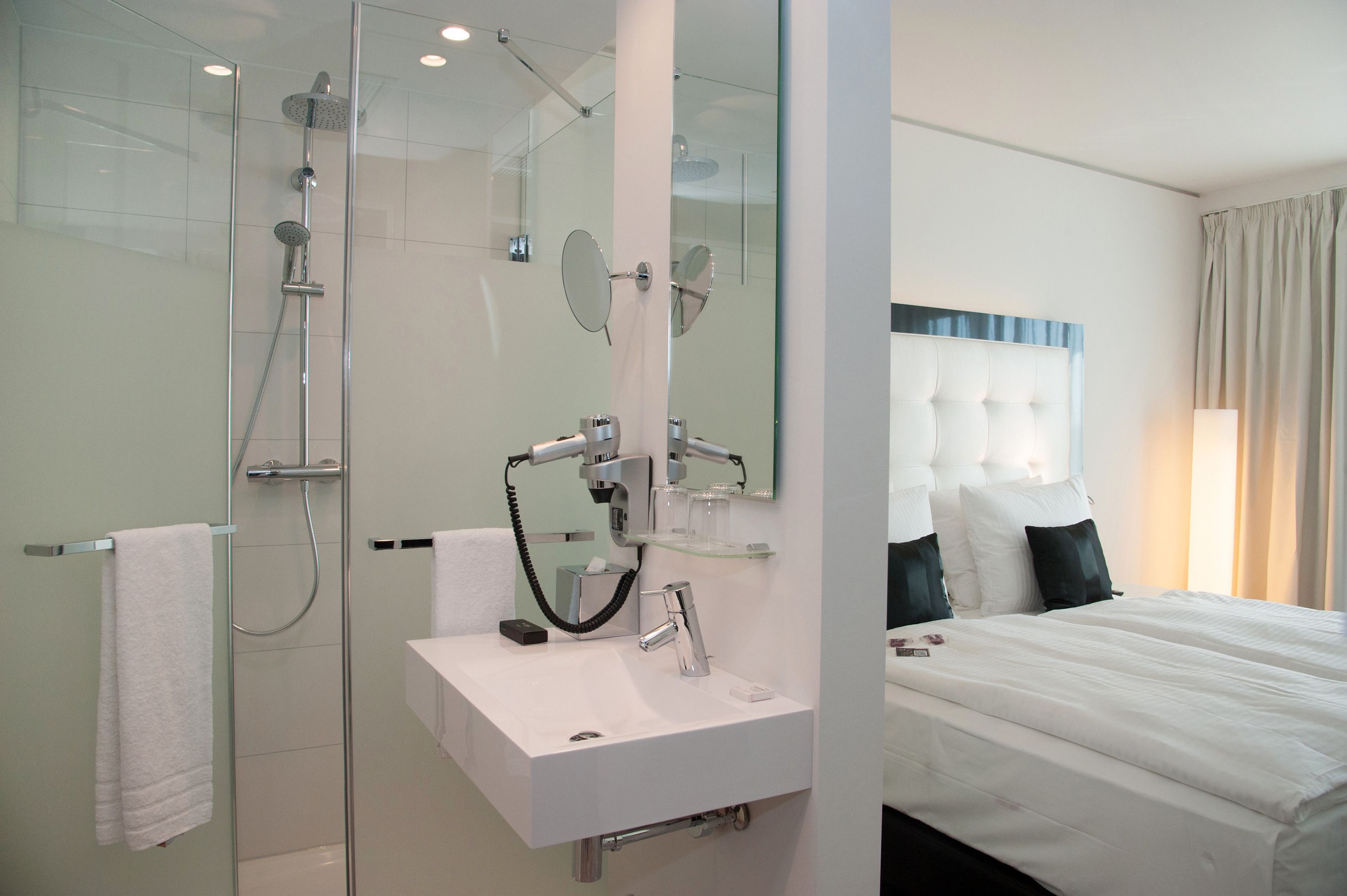 A modern bathroom with a glass-enclosed shower, a white sink with a mirror, and a bed with white bedding and dark pillows.