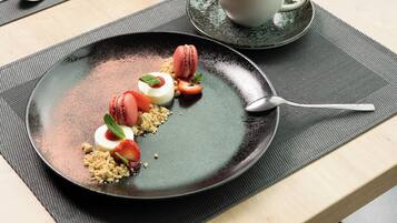 A dessert plate with macarons, strawberries, and a mint garnish on a dark plate, accompanied by a cup of coffee on a saucer.