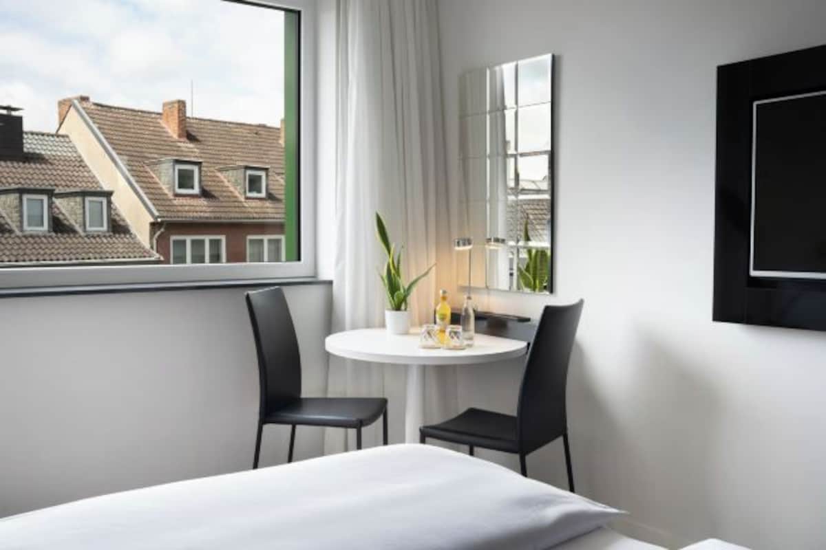 Habitación de hotel con cama, una mesa redonda con dos sillas, una planta en maceta y un ventanal con vistas a edificios cercanos.
