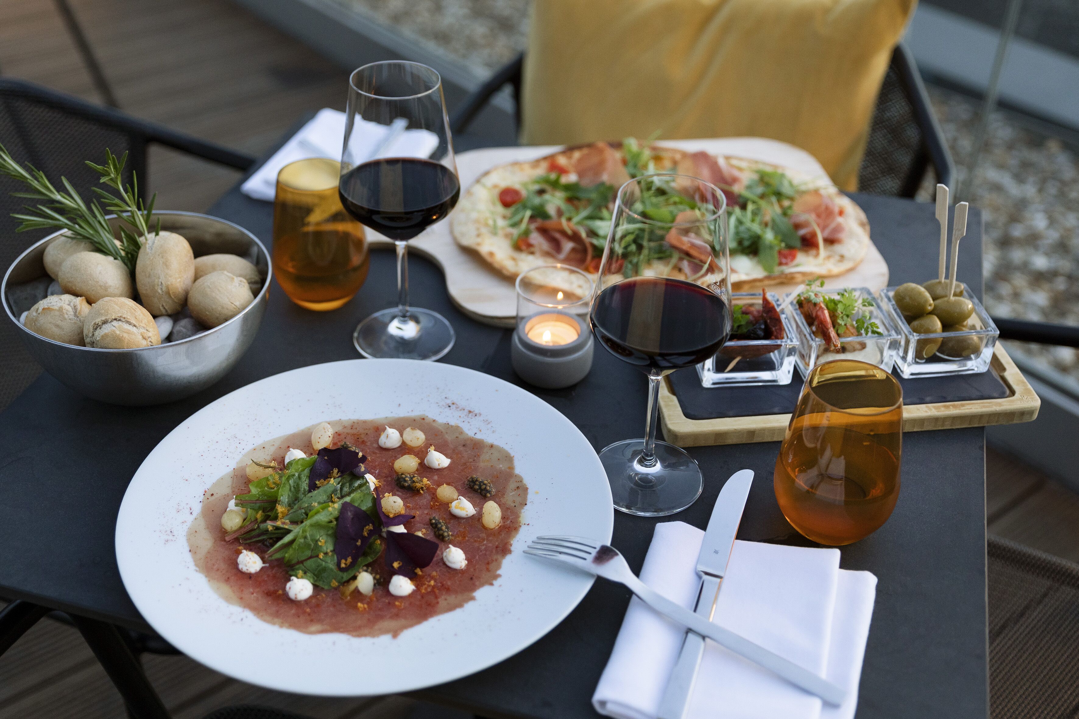 A table set with a variety of dishes, including a pizza, a salad, and a bowl of olives, accompanied by glasses of wine.