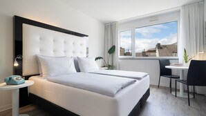 Una habitación de hotel moderna con una cama grande, una mesita de noche con lámpara, un escritorio pequeño con silla y una ventana con vista a la ciudad.