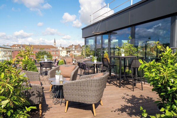 Una terraza en la azotea con mobiliario de mimbre, barandilla de vidrio y vistas de la ciudad.