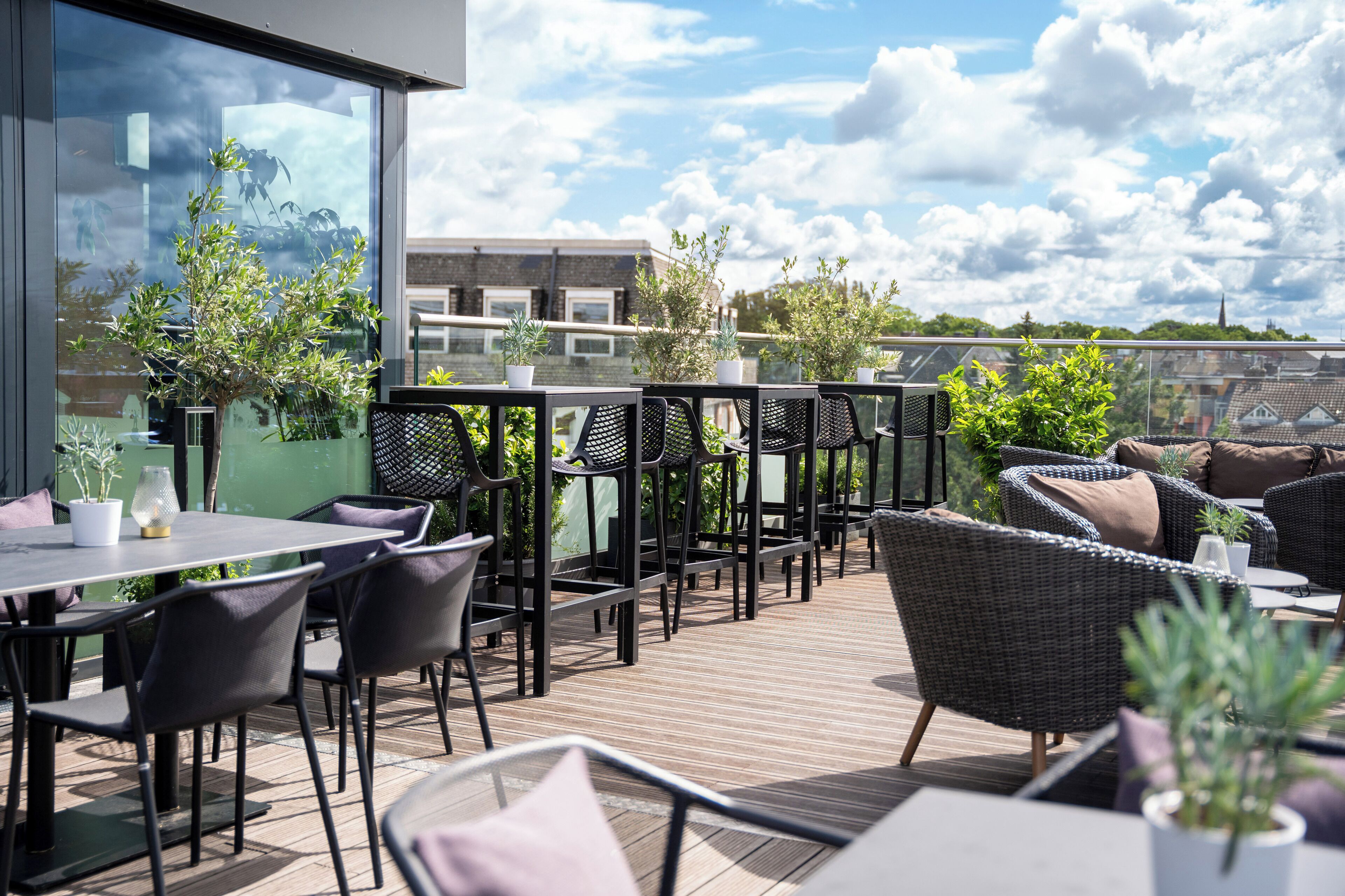 Um terraço no telhado com mesas ao ar livre, plantas em vasos e vista para edifícios e céu.