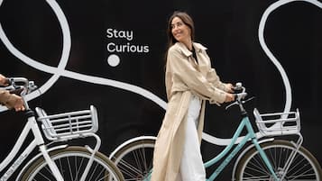 A person riding a turquoise bicycle with a white basket in front, standing next to a black wall with white text and graphics.