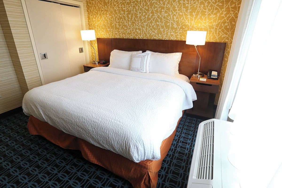 A hotel room with a large bed, two bedside lamps, a nightstand, and a wall-mounted air conditioner.