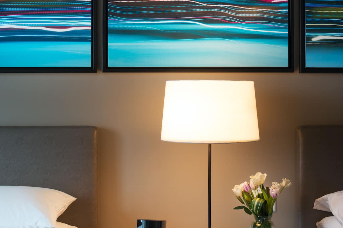 Quarto de hotel com uma cama, mesa de cabeceira com abajur, vaso com flores e três obras de arte emolduradas na parede.