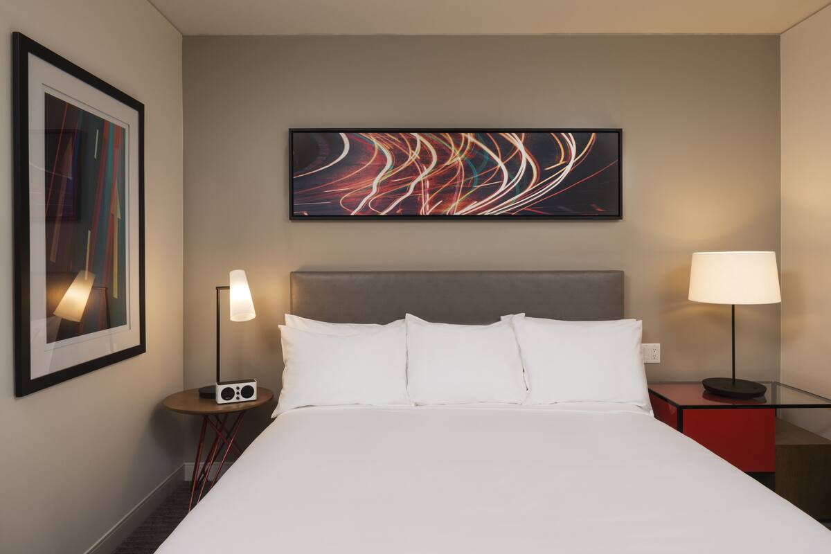 Quarto de hotel com uma cama grande, duas mesas de cabeceira com luminárias, um quadro na parede e uma obra de arte abstrata sobre a cama.