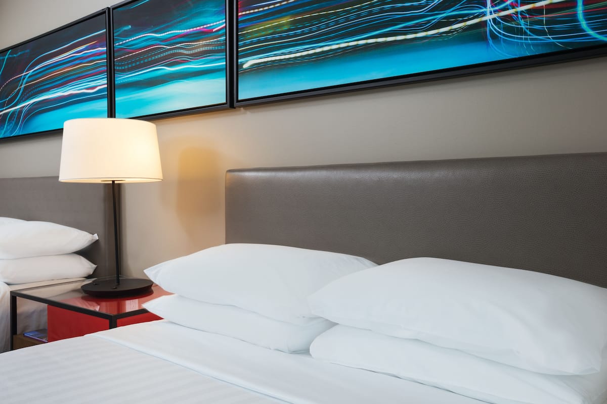 Quarto de hotel com cama, almofadas brancas, mesa de cabeceira e luminária, além de arte abstrata nas paredes.