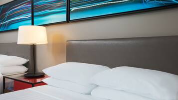 Hotelkamer met een bed, witte kussens, een nachtkastje met een lamp en abstracte wanddecoratie.