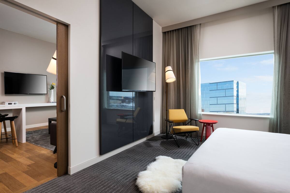 Quarto de hotel moderno com uma cama grande, uma escrivaninha com televisão de tela plana, uma cadeira, uma mesinha e vista para um edifício pela janela.