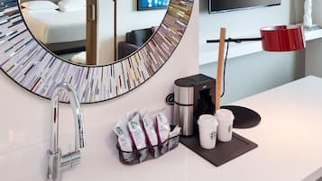 A modern bathroom with a large mirror, a sink, a coffee maker, and a flat-screen TV on the wall.