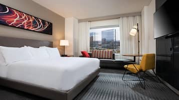 A hotel room with a large bed, a desk, a chair, a TV, and a view of buildings through the window.