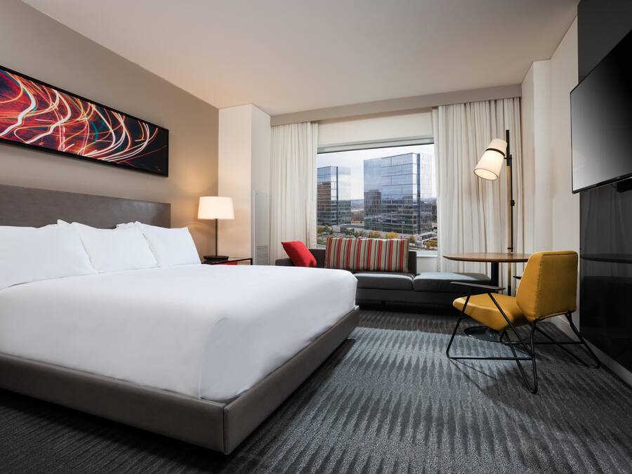 A hotel room with a large bed, a desk, a chair, a TV, and a view of buildings through the window.