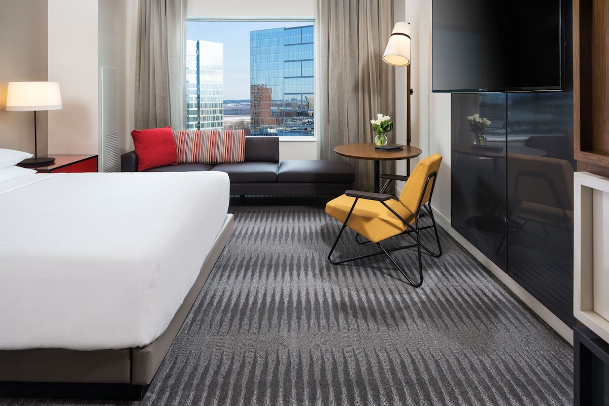 Quarto de hotel com uma cama grande, um sofá, uma mesinha com um vaso, uma televisão e vista para a cidade.