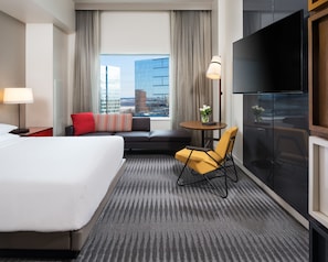Quarto de hotel com uma cama grande, um sofá, uma mesinha com um vaso, uma televisão e vista para a cidade.