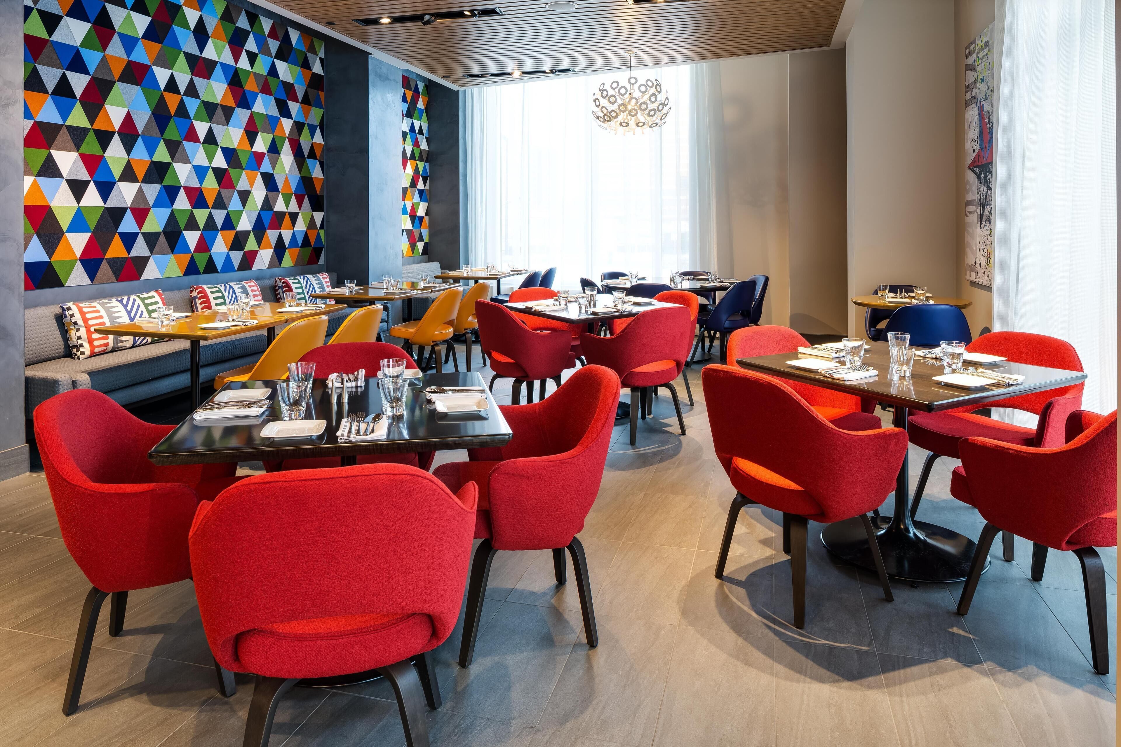 A modern restaurant interior with red and orange chairs, wooden tables, and a colorful geometric wall design.