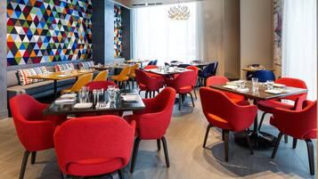 Un intérieur de restaurant moderne avec des chaises rouges et orange, des tables en bois et un mur décoré de motifs géométriques colorés.