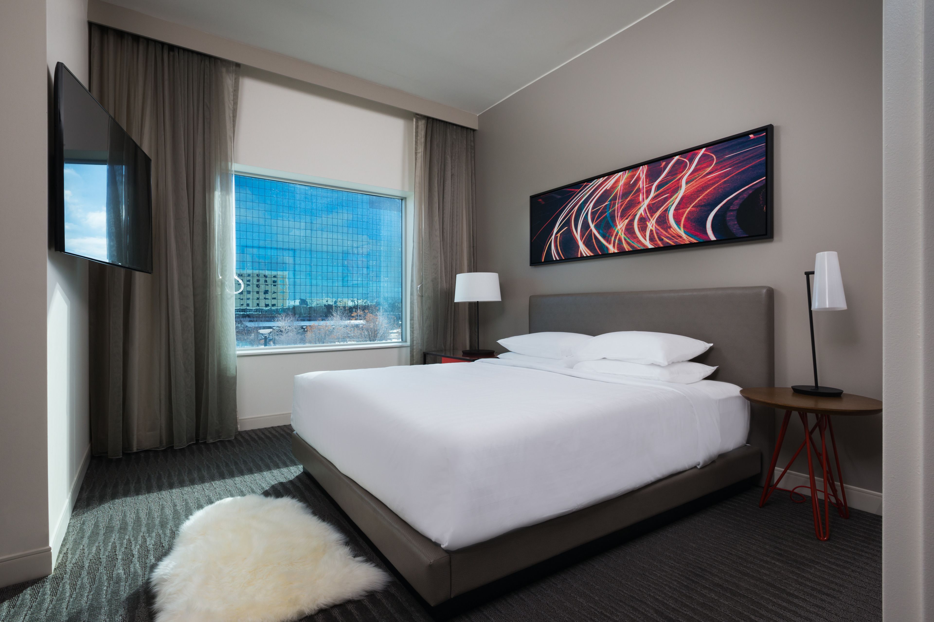 A hotel room with a large bed, a flat-screen TV, a window with a city view, a modern abstract painting, and a small table with a lamp.