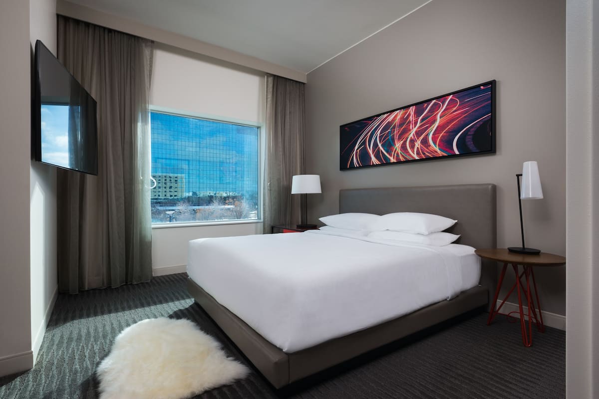 Quarto de hotel com uma cama grande, televisão de tela plana, janela com vista para a cidade, pintura abstrata moderna e uma pequena mesa com luminária.