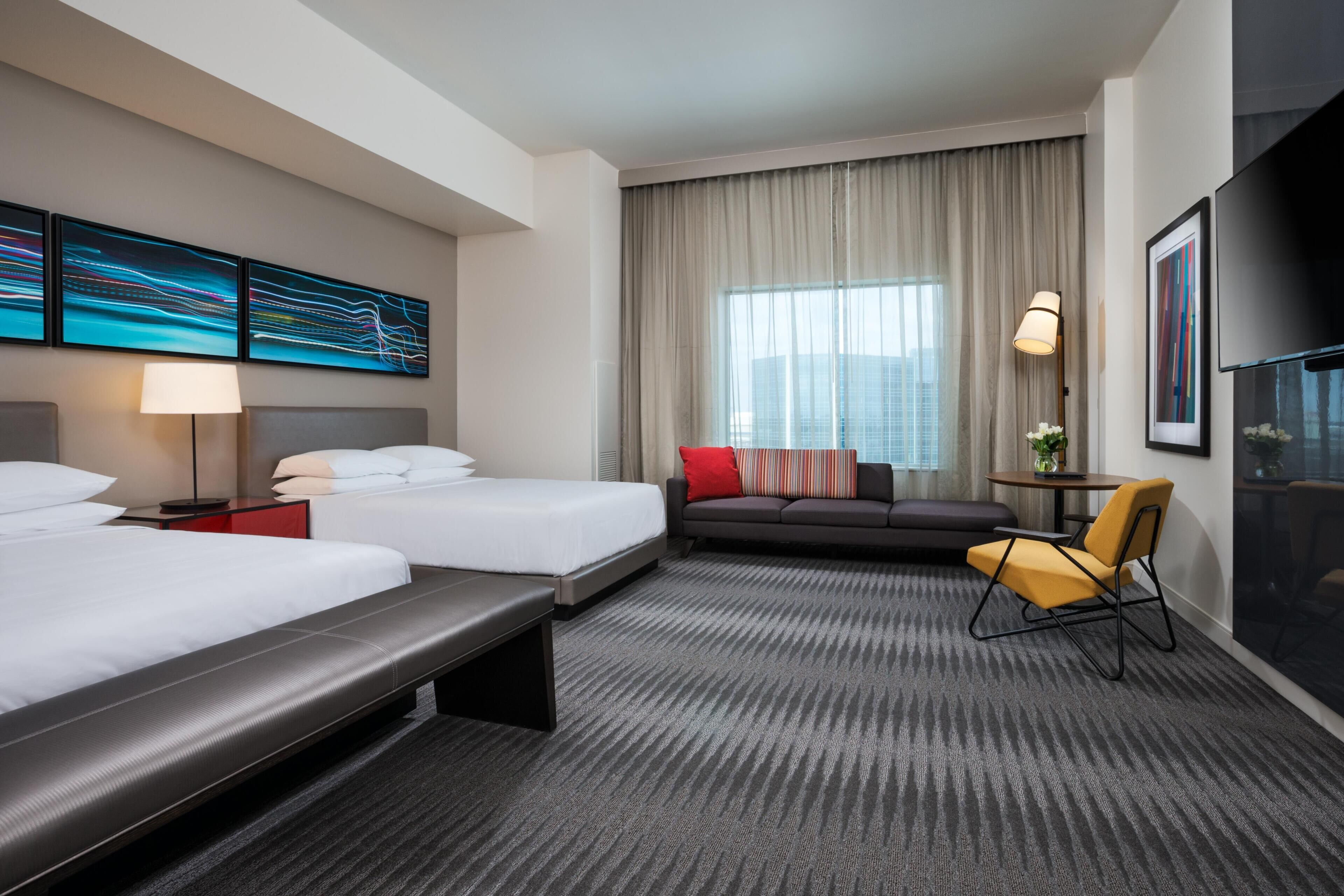 Habitación de hotel con dos camas, un sofá, una silla, un escritorio y un ventanal con cortinas.