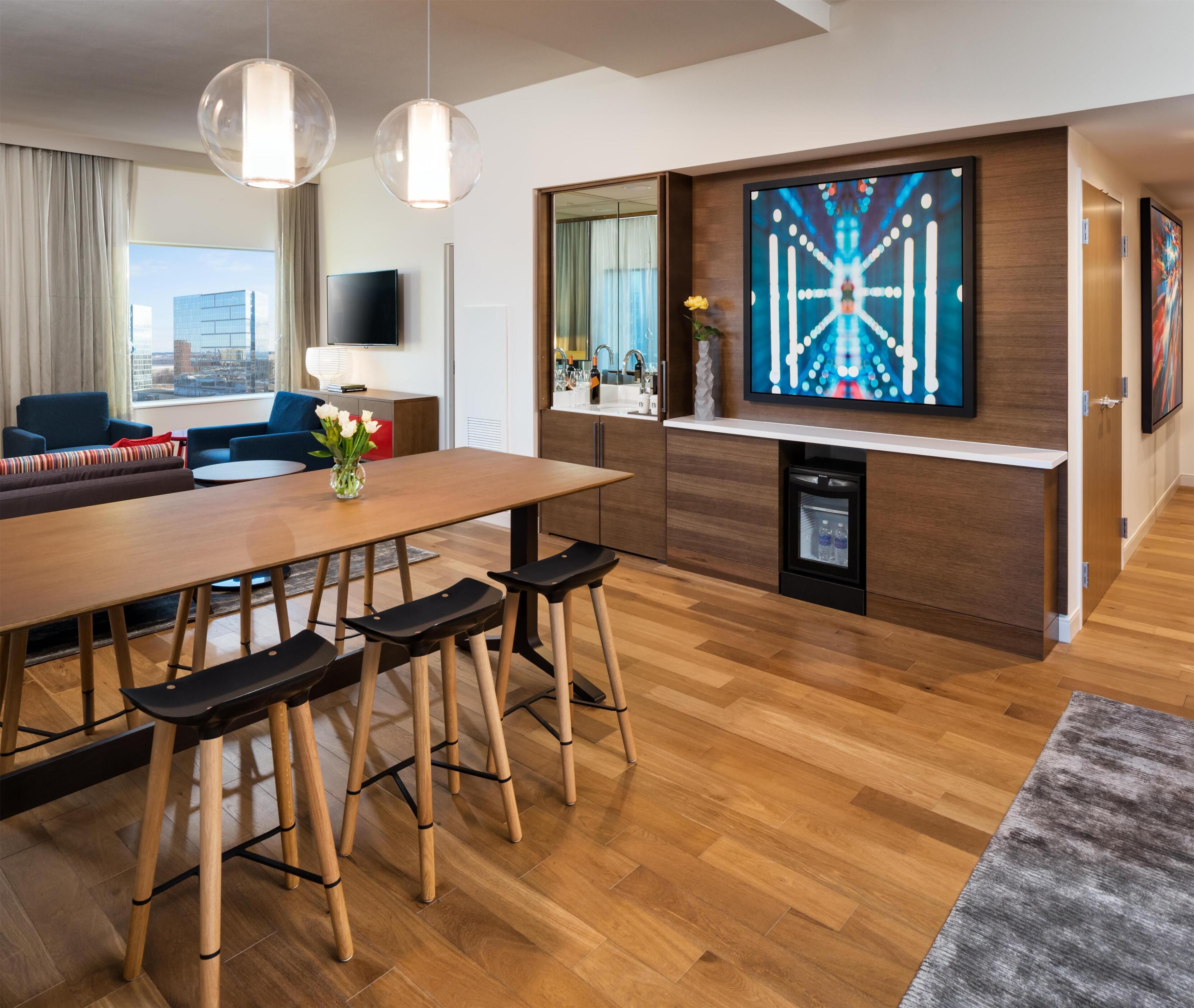 A modern hotel room with a wooden dining table, bar stools, a flat-screen TV, a built-in sink, and a city view.