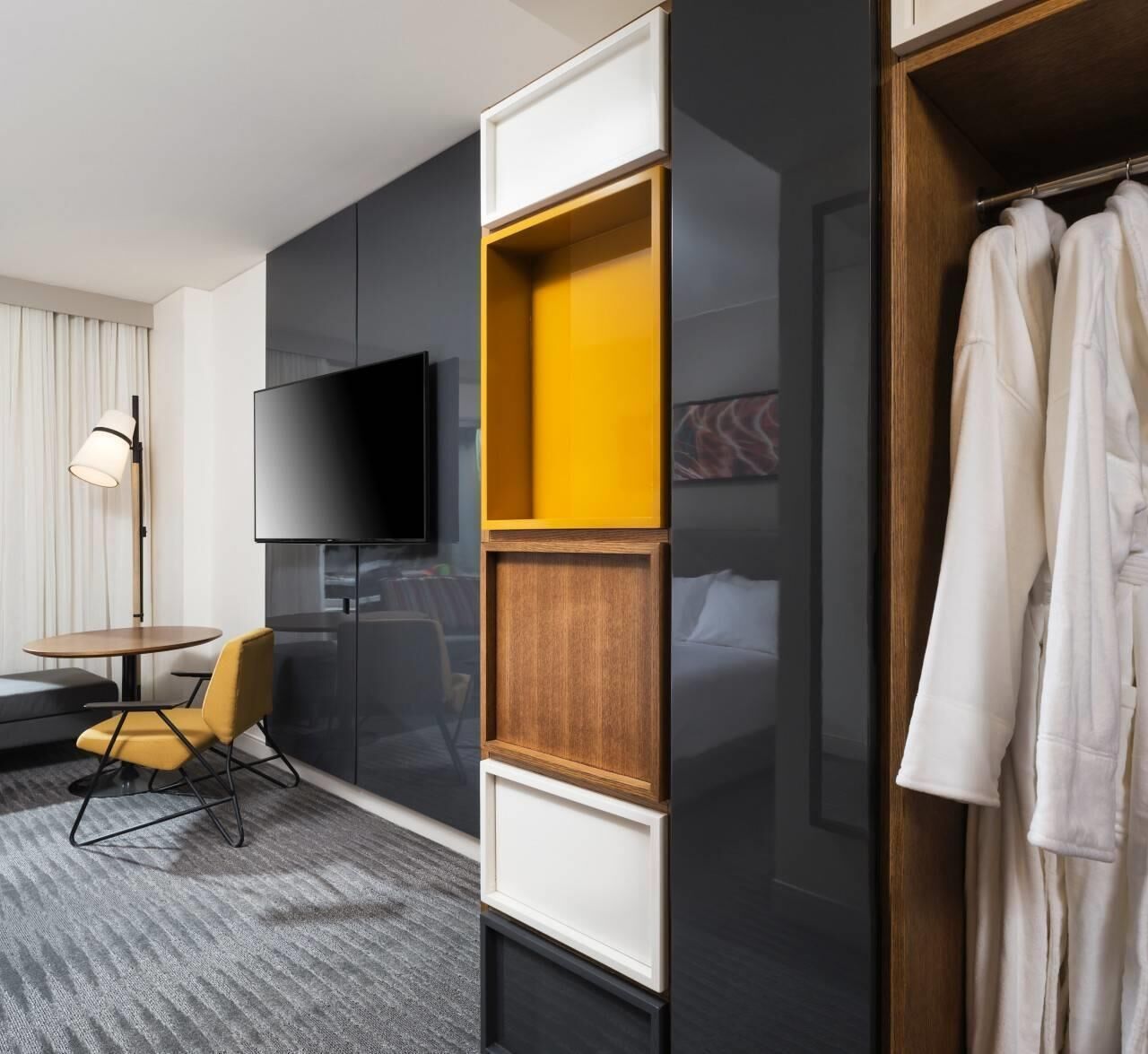 Habitación de hotel moderna con una televisión de pantalla plana, una mesita redonda, una silla amarilla y un armario con espejo.