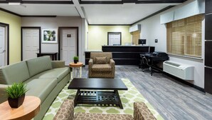Una sala moderna con un sofá, sillón, mesa de centro y una mesita auxiliar con una planta. Se ve una cocina con encimera y electrodomésticos al fondo.